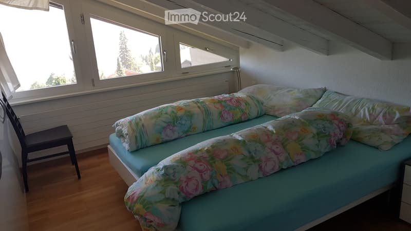 Cozy bedroom with a single bed covered in a floral duvet, a small wooden chair, and large windows offering a view of trees and a house outside.