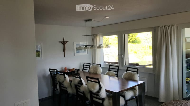 Dining room with a wooden dining table surrounded by chairs with fur cushions, large windows with curtains, and a cross on the wall.