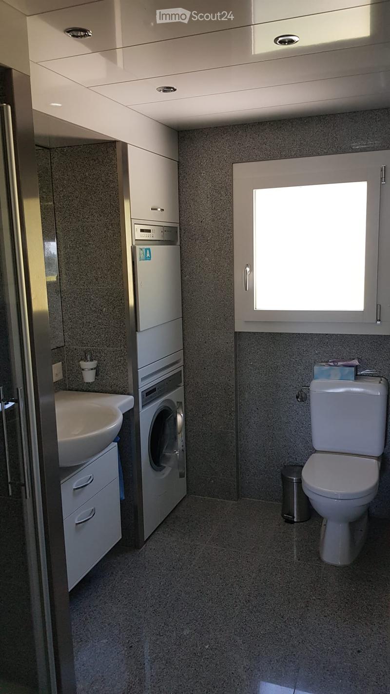 Modern bathroom with a sink, toilet, washing machine, and built-in cabinets. Features a window and a small trash can.