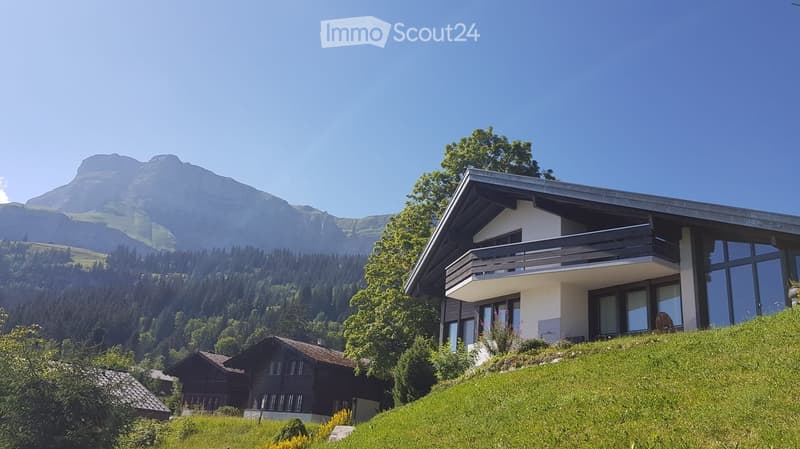 Modern two-story house with large windows and a balcony, situated on a grassy slope with a mountain view in the background.