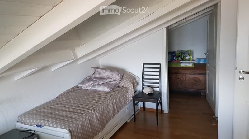Cozy attic bedroom with a single bed, a small chair, and a stool holding a hat. The room features slanted wooden ceilings and white walls.