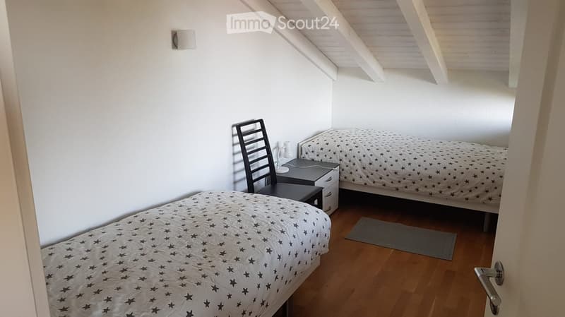 Cozy attic bedroom with two single beds, a nightstand, a chair, and a lamp. The room features wooden flooring and white walls with a slanted ceiling.