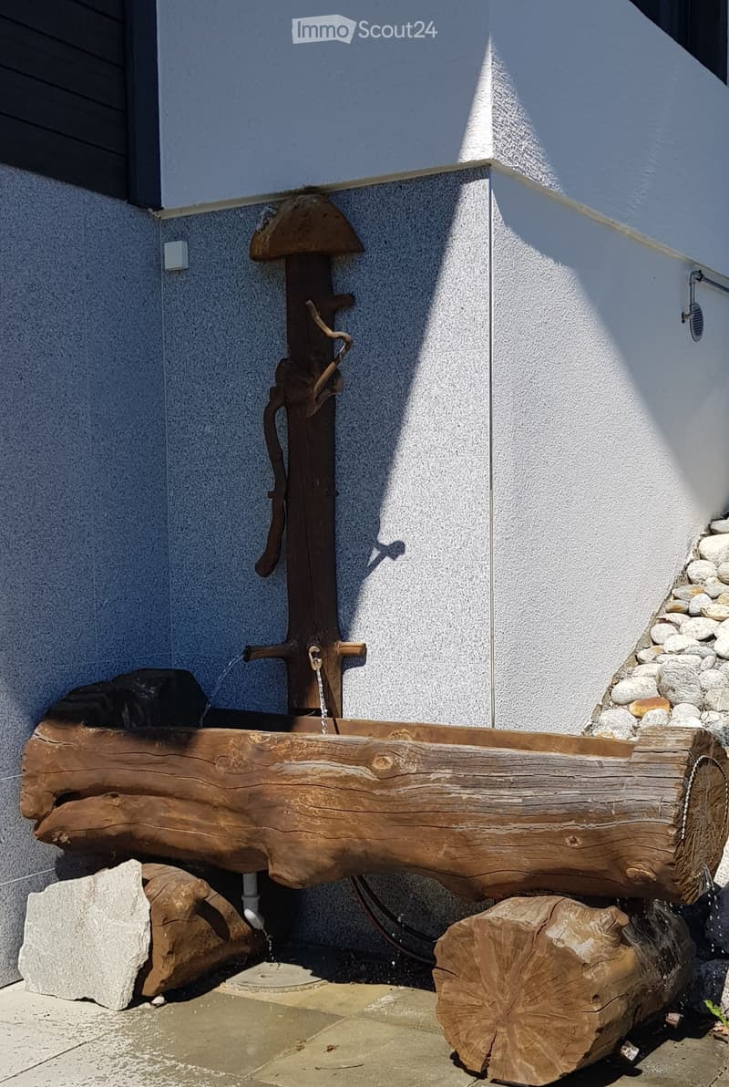 A rustic wooden water pump with a log-shaped basin, situated against a modern building wall with stone accents and a small staircase.
