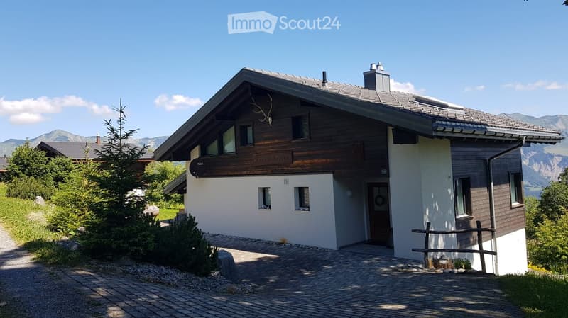 Two-story house with a wooden facade, white walls, and a gray roof, surrounded by greenery and mountains.