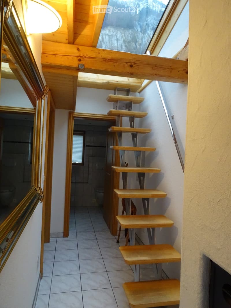 Wooden staircase with multiple steps leading upwards, white tiled floor, and a view of a bathroom area with a toilet and shower stall visible in the background.