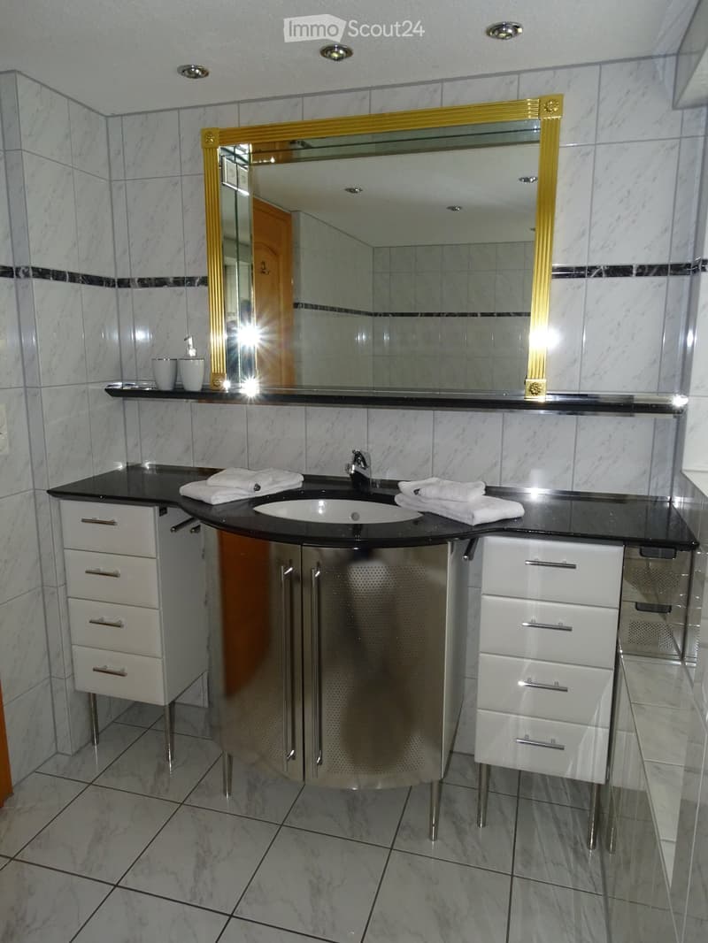 Modern bathroom with marble tiles, a large sink, a mirror with gold frame, and white cabinets.
