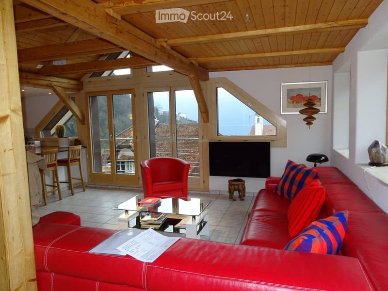 Living room with exposed wooden beams, large windows, red leather sofa, glass coffee table, red armchair, small wooden stool, and a dining area with a table and chairs.