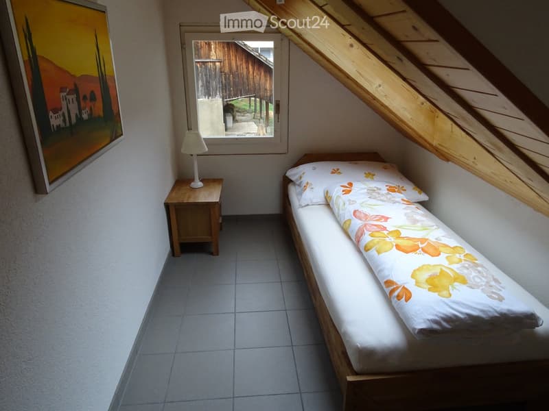 Cozy attic bedroom with a single bed, nightstand, lamp, and a painting on the wall. The room features slanted wooden ceilings and a window offering a view of the outdoors.