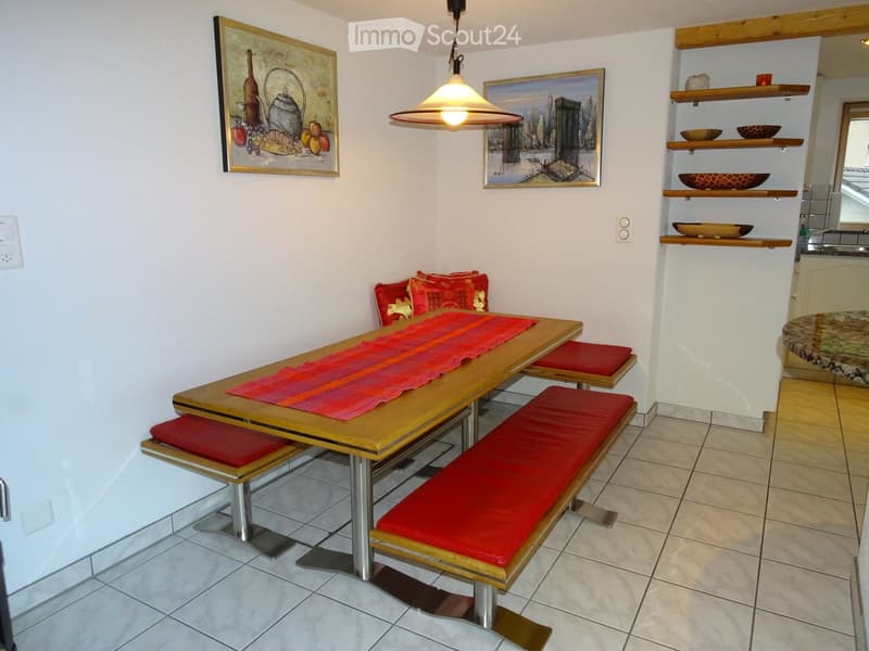 Cozy kitchen area with a wooden dining table, red cushioned benches, pendant light, shelves with decorative items, and framed artwork on the walls.