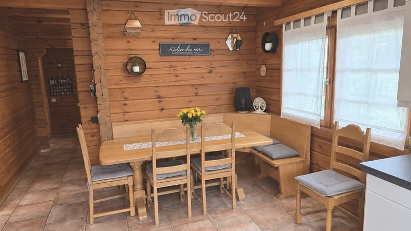 Cozy wooden dining area with a round table, four chairs, a bench with cushions, and a vase of yellow flowers. Wooden walls and tiled floor.