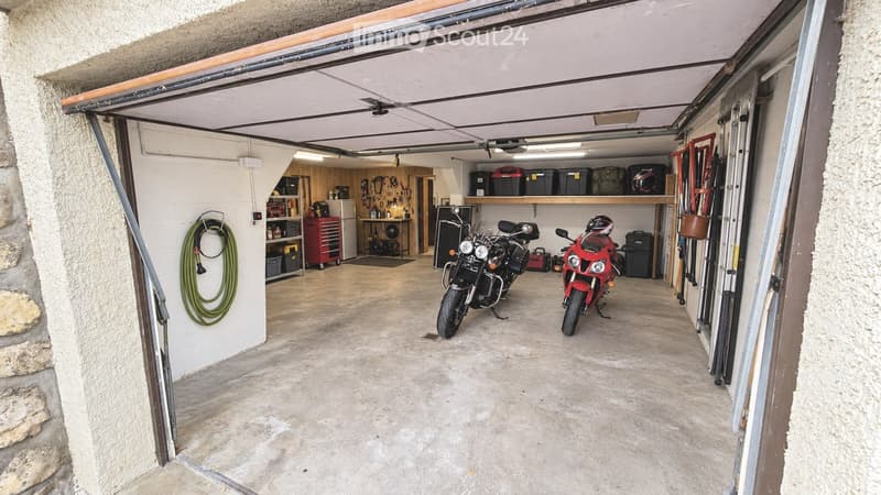 Garage with two motorcycles, storage shelves, and tools. Garage door is open, revealing a concrete floor and organized storage area.