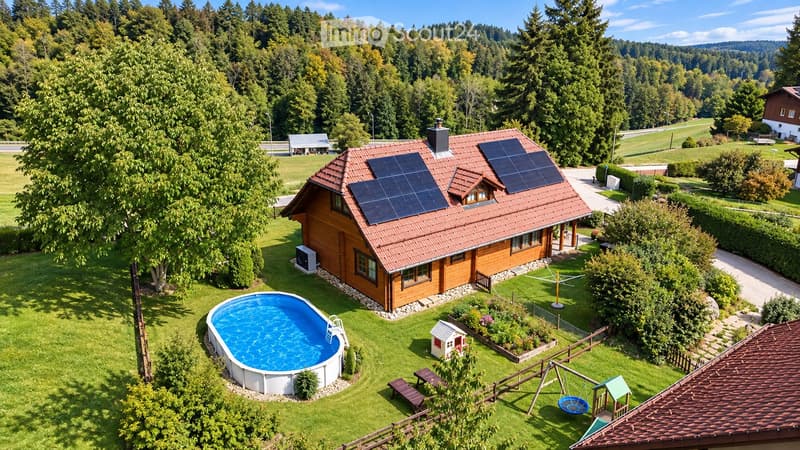 Aerial view of a wooden house with solar panels on the roof, surrounded by a well-maintained garden with a swimming pool, a small playhouse, and a swing set.