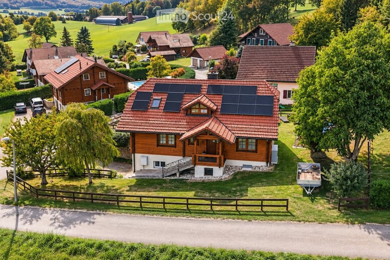 Aerial view of a residential area with multiple houses, solar panels on roofs, a swimming pool, and surrounding greenery.
