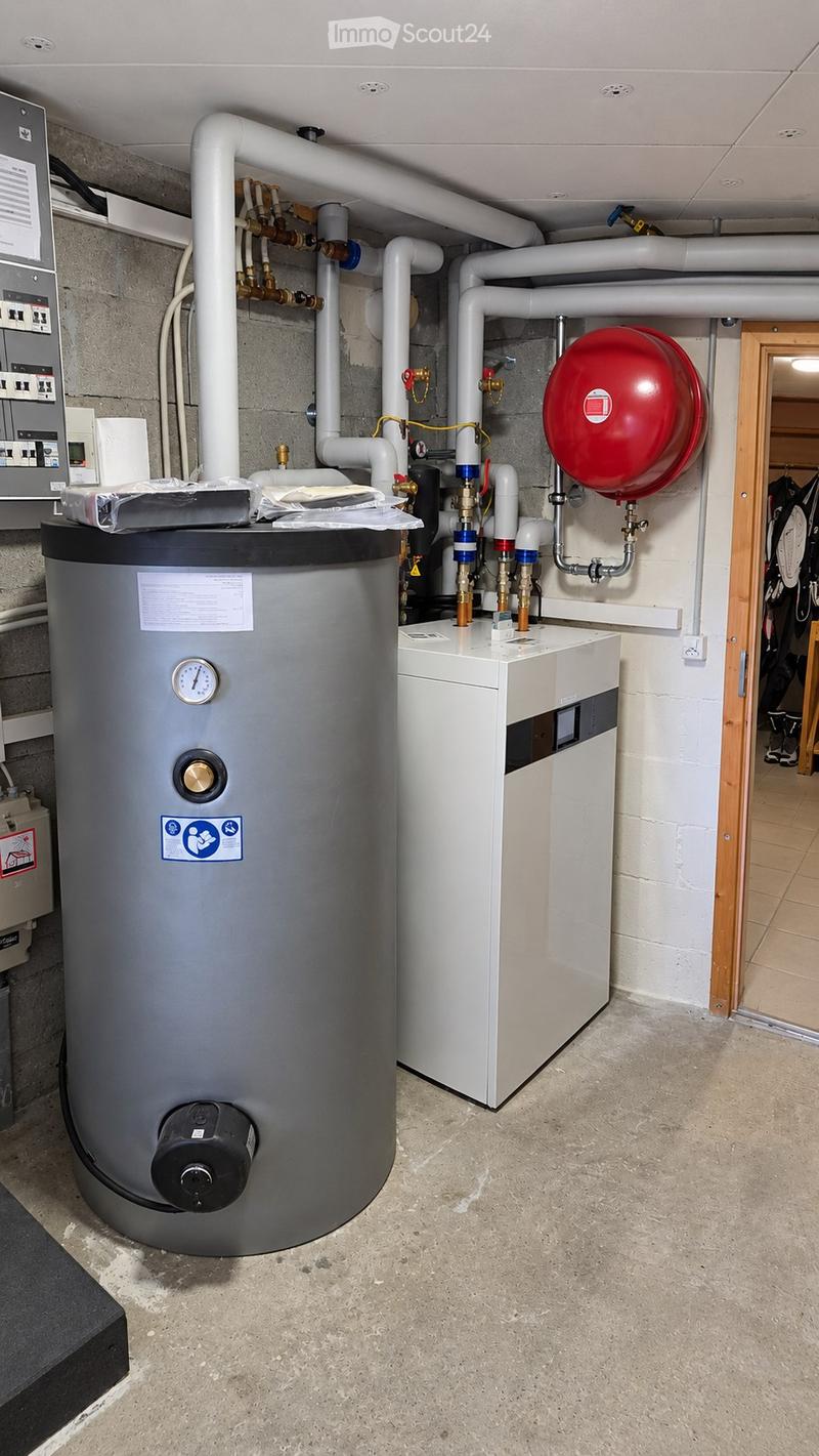 Utility room with a water heater, boiler, and various pipes and valves. The room has a concrete floor and exposed brick walls.