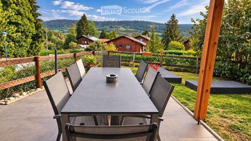 Outdoor patio with a black table and six chairs, surrounded by a wooden fence, lush greenery, and a scenic mountain view.