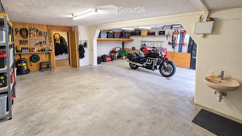 Garage with concrete floor, wooden wall, shelves, tools, motorcycle, lawn mower, sink, and storage bins.