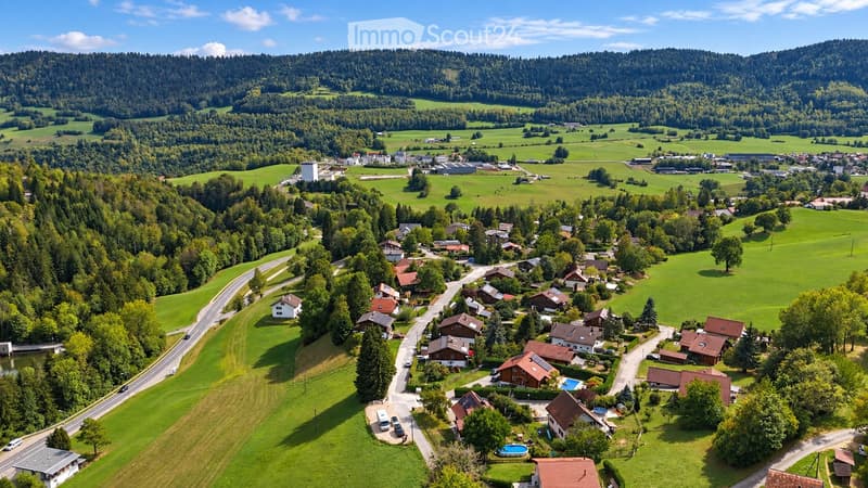 Aerial view of a rural village nestled in a lush green valley surrounded by forested hills. The village features scattered houses with red-tiled roofs, surrounded by fields and trees. A small swimming pool is visible in one of the yards.