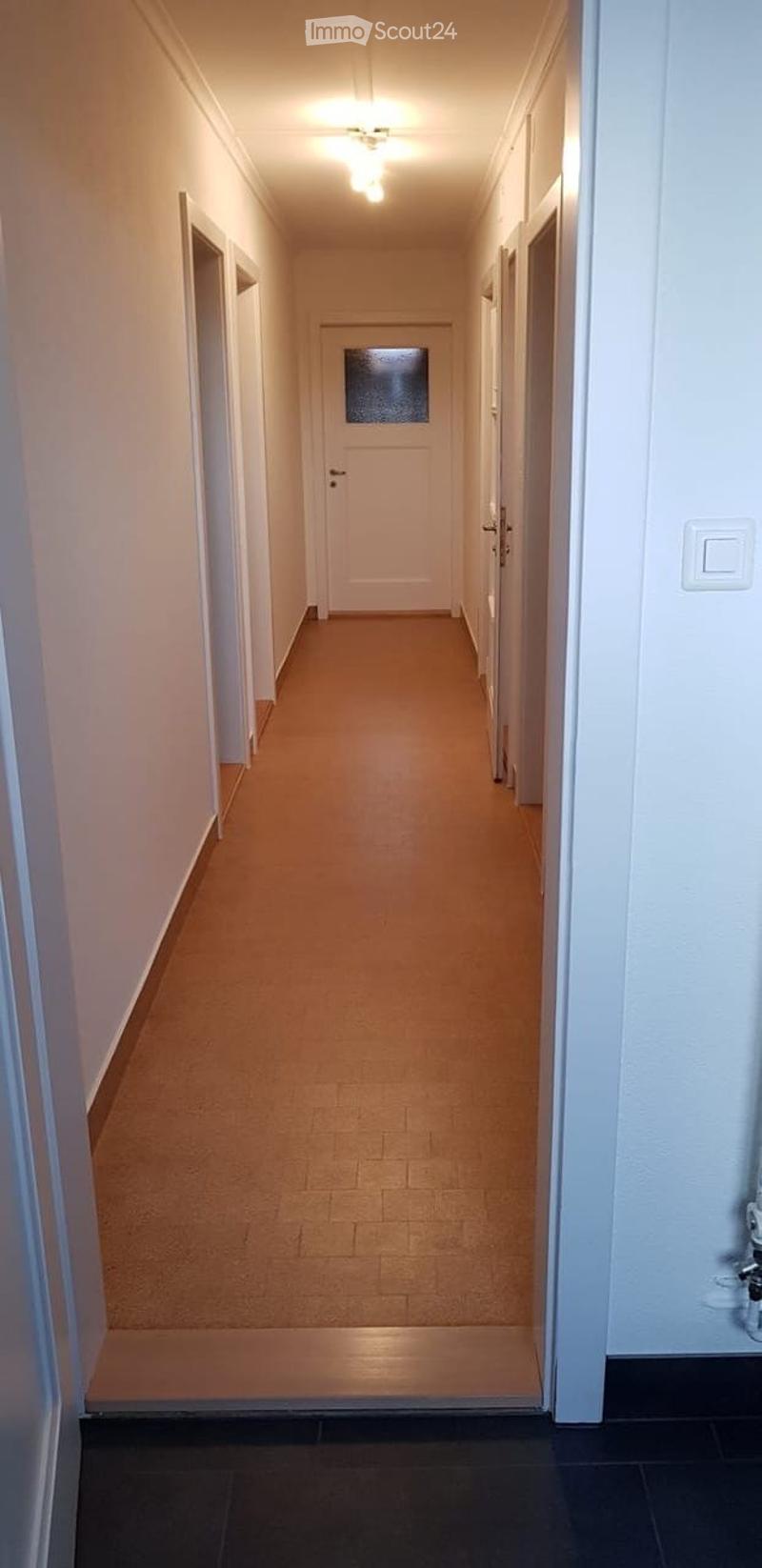 A long, narrow hallway with light-colored walls, a beige tiled floor, and white doors on both sides. A ceiling light fixture provides illumination.