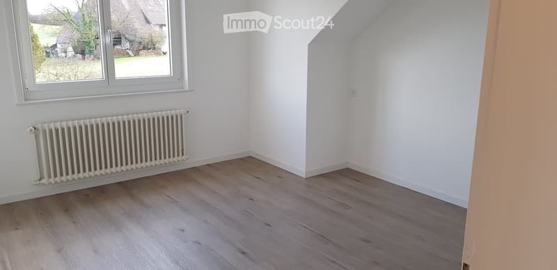 Empty room with wooden flooring, white walls, a radiator, and a large window offering a view of the outdoors.