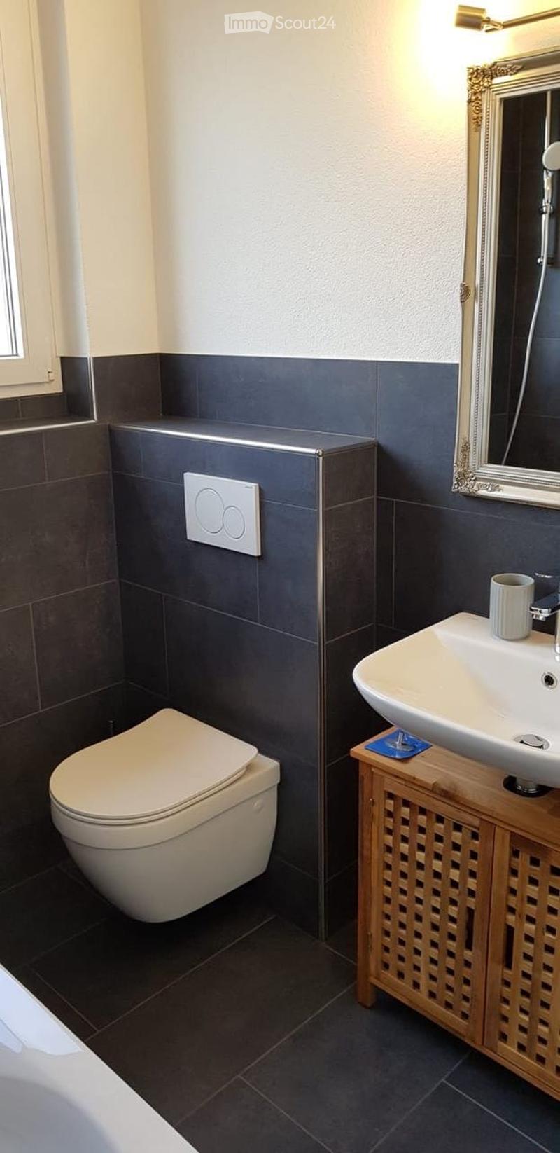 Modern bathroom with dark grey tiles, white toilet, white sink, wooden cabinet, and a decorative mirror.