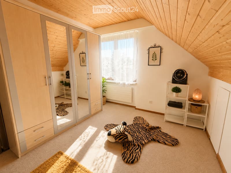Bright bedroom with wooden slanted ceiling, large window with sheer curtains, wardrobe with mirrored doors, small shelf with plant and decorative items, and a large stuffed animal on the floor.