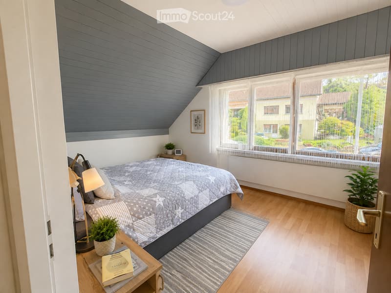 Cozy attic bedroom with wooden floors, slanted grey ceiling, large windows with white curtains, bed with grey and white bedding, nightstand with lamp, side table with books and plant, and a small potted plant on the floor.