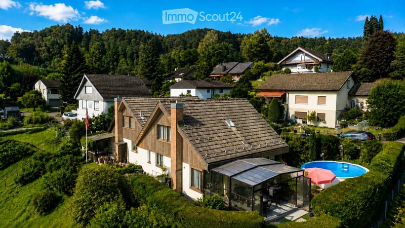 Aerial view of a modern house with a tiled roof, surrounded by greenery and neighboring houses. Features include a swimming pool, patio area, and outdoor furniture.