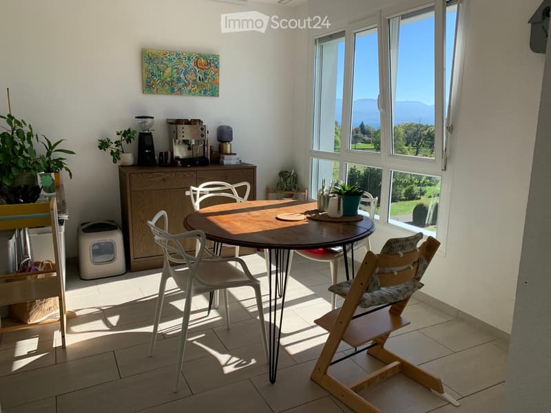 Bright dining area with a round wooden table, white chairs, a baby chair, a wooden cabinet with a coffee machine, and potted plants. Large windows offer a scenic view.