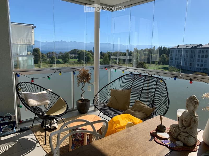 Balcony with a view of mountains and water, featuring a wicker chair, a sofa, a small table, a potted plant, and a decorative statue.