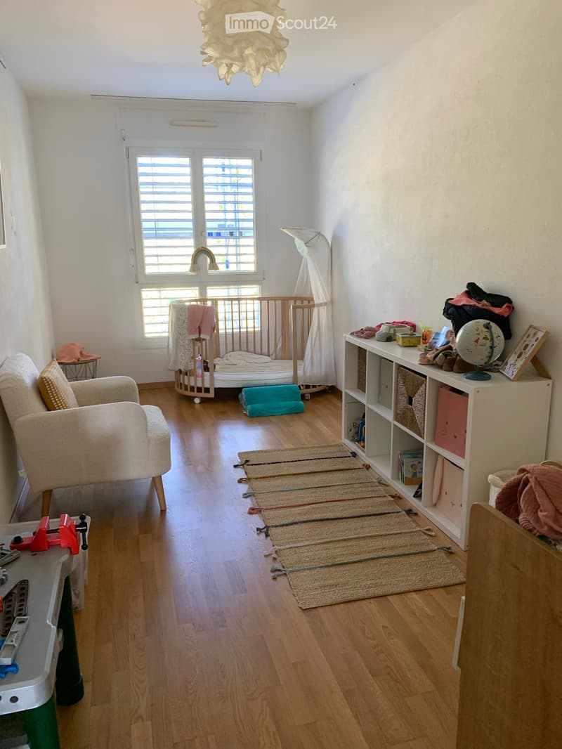 A well-lit children's room with wooden flooring, a baby crib, a shelf with toys and books, a comfortable chair, and a small table with a globe and picture frame.