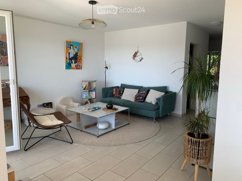 Modern living room with a teal sofa, white coffee table, wicker chair, and decorative plant in a woven pot. The space features a round rug, a bookshelf, and a large mirror on the wall.