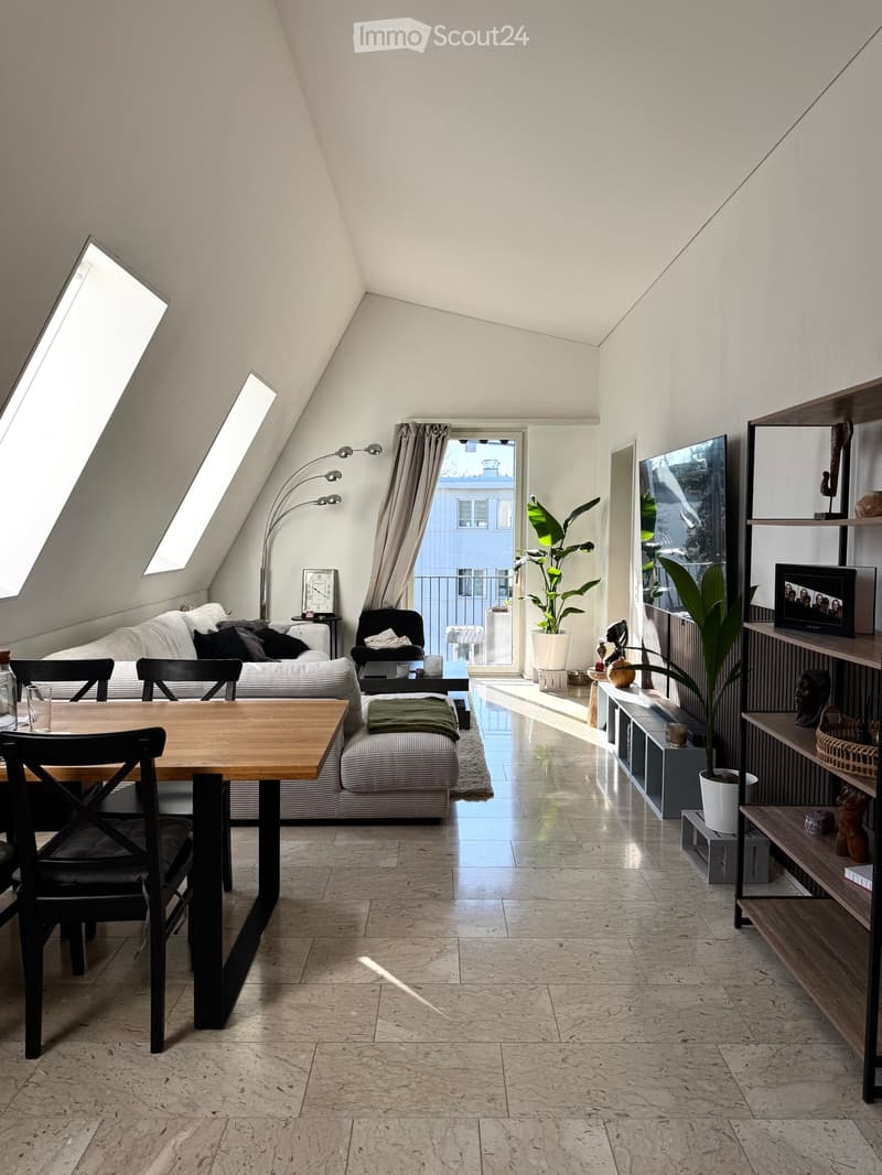 Bright, modern living space with a slanted ceiling, large windows, and a balcony. Features a sofa, dining table, chairs, shelves, a floor lamp, and plants.