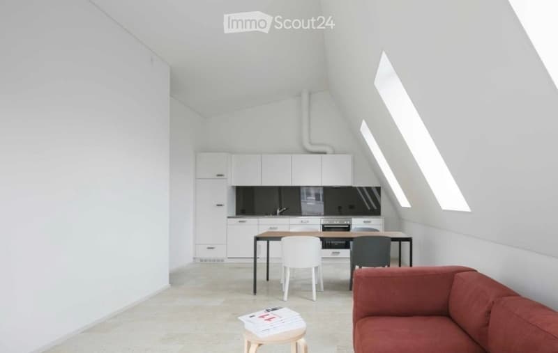Modern, minimalist living and dining area with white walls, a red sofa, a wooden dining table with chairs, a small stool with magazines, and a sleek white kitchen with cabinets and an oven.