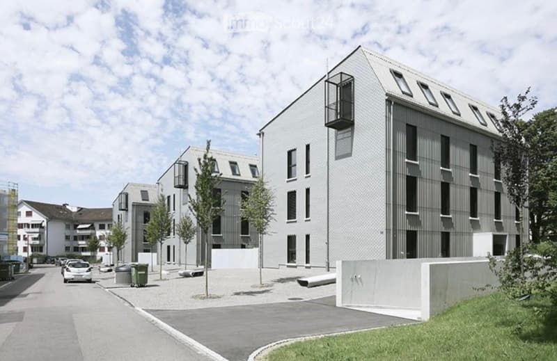 Modern multi-story building with a sleek, contemporary design, featuring large windows and a flat roof. The building is surrounded by a paved area with a few trees and a parked car.