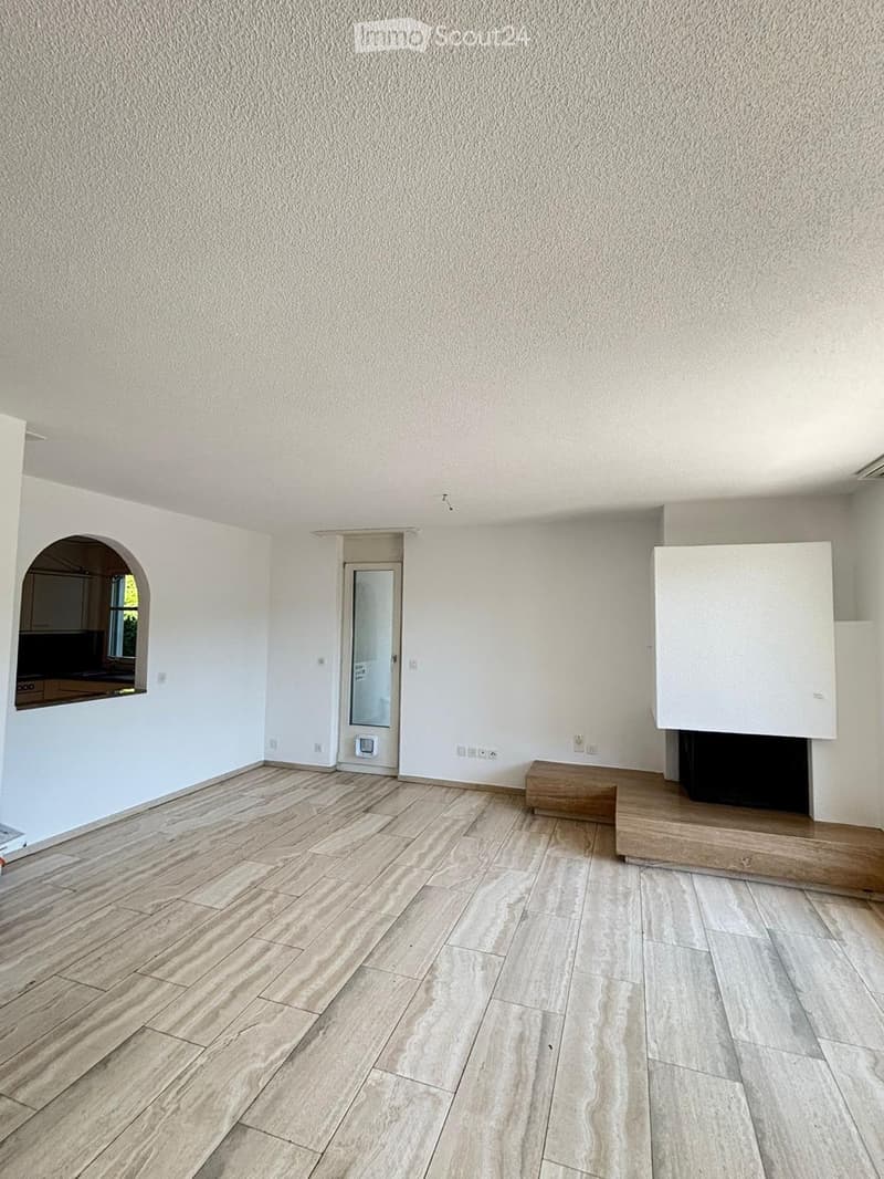 Empty room with light-colored walls, wooden flooring, and a fireplace with a wooden bench in front of it. There is a doorway leading to another room and an arched opening to a kitchen area.
