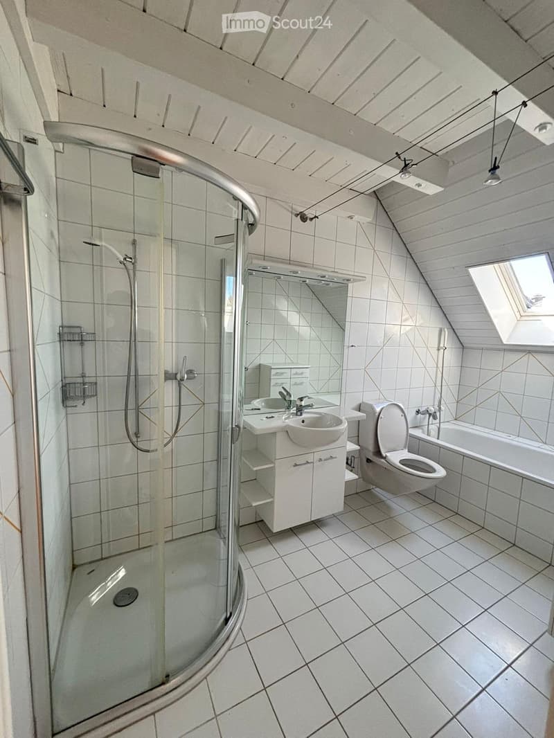 Modern bathroom with white tiled walls and floor, featuring a glass-enclosed shower, a sink with a mirror, a toilet, and a bathtub. The ceiling has exposed wooden beams and a skylight window.