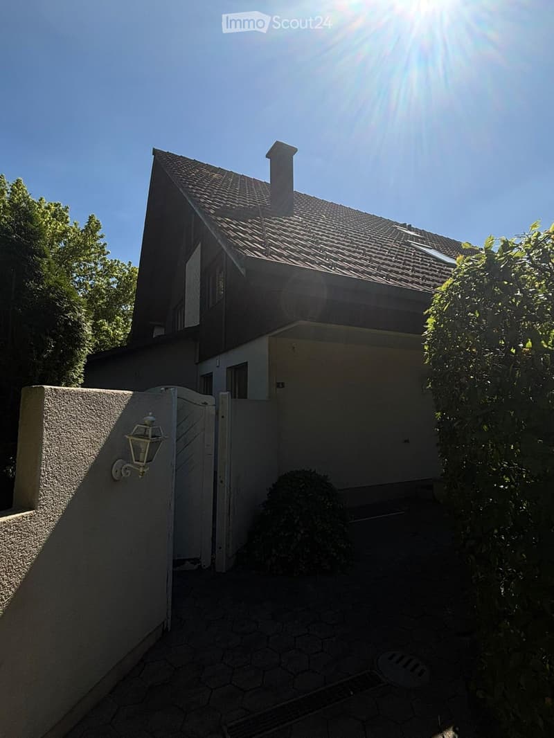 Two-story house with a tiled roof, chimney, and garage. The entrance features a white gate and a wall-mounted lantern. The surrounding area includes a paved pathway and greenery.