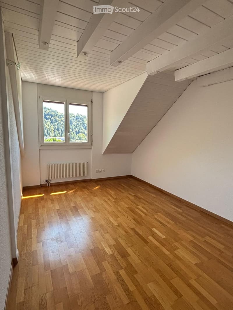 Empty room with wooden flooring, white walls, and a slanted ceiling. Features include a window with a view of greenery, a radiator, and a door.