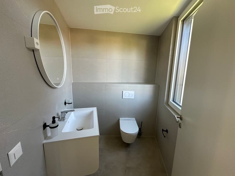 Modern bathroom with a white sink, faucet, toilet, and a round mirror on the wall. The room features light grey tiles and a window providing natural light.