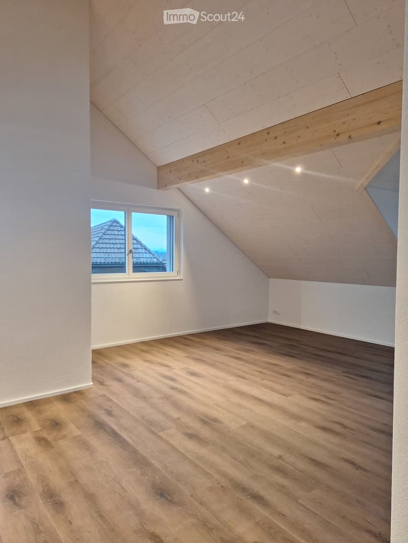 Empty attic room with wooden flooring, white walls, and a single window offering a view of the roof.