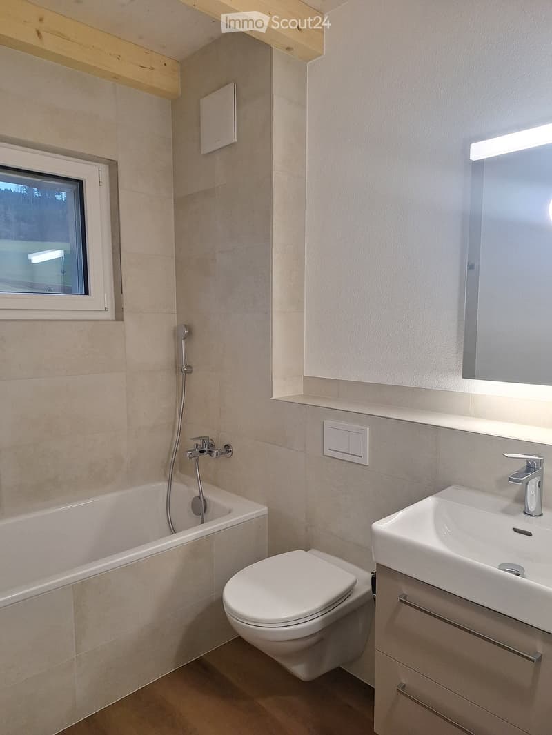 Modern bathroom with a bathtub, toilet, sink, and mirror, featuring beige tiles and a window with a view.