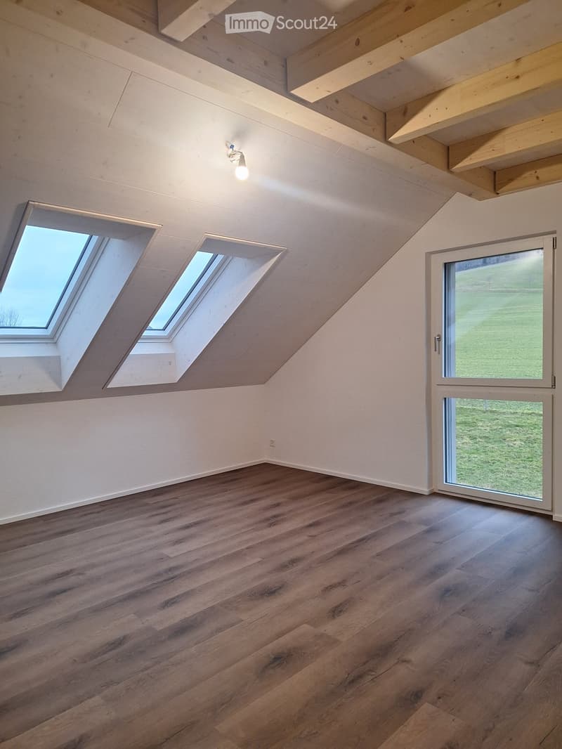 Empty room with wooden flooring, white walls, exposed wooden beams on the ceiling, and skylight windows. A sliding glass door leads to an outdoor area.