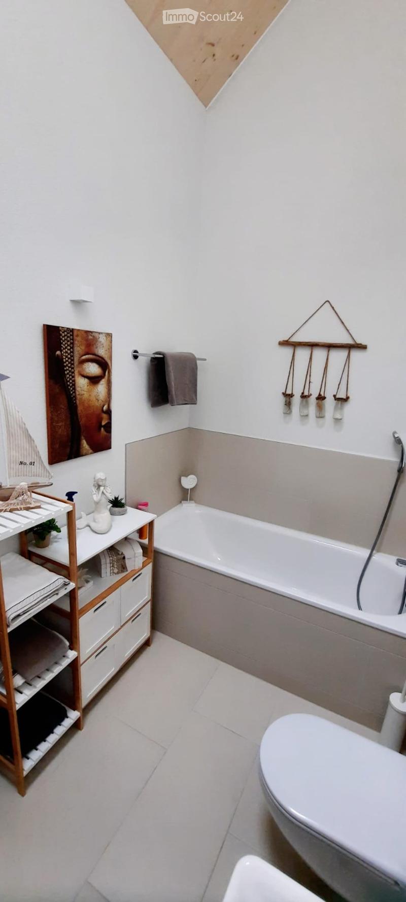 Modern bathroom with white tiles, a bathtub, toilet, wooden shelf with towels and decorative items, and a towel rack with towels.