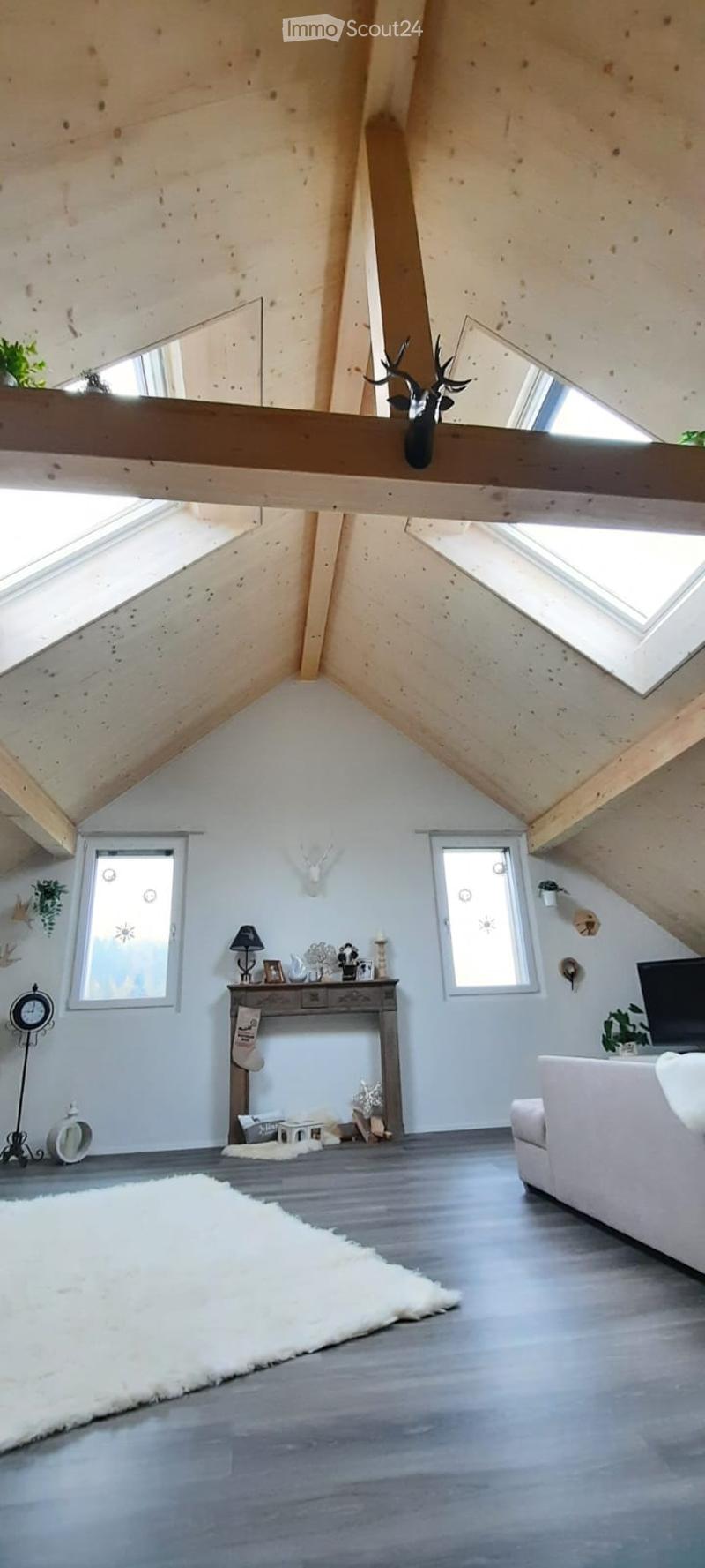 Bright attic living space with wooden beams, skylights, and large windows. Features a white sofa, wooden coffee table, floor lamp, and decorative items.