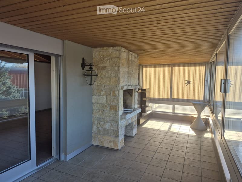 Balcony with tiled floor, stone fireplace, white stool, and large windows with blinds.
