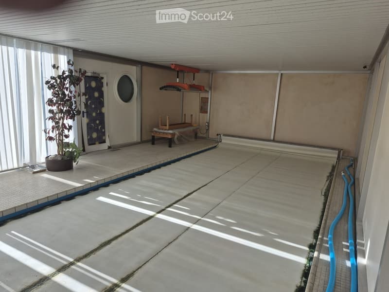 A covered outdoor area with a tiled floor, a small wooden stool, a potted plant, and a door with a round window.