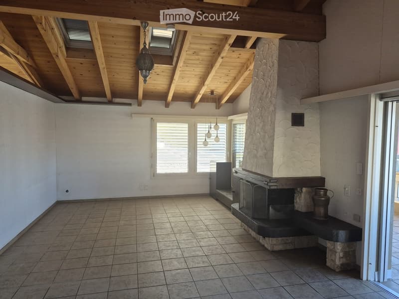 Living room with exposed wooden ceiling beams, large windows, and a stone fireplace with a decorative metal vase on the mantel.