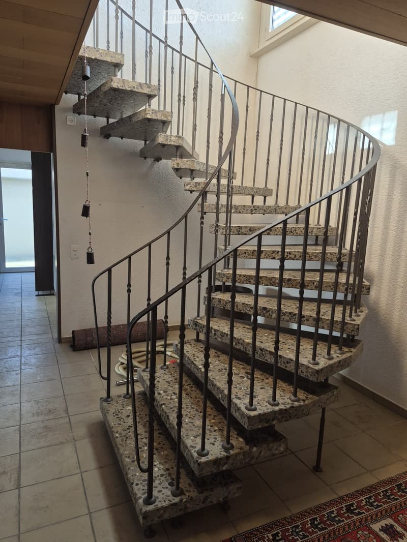 Spiral staircase with stone steps and metal railing, leading to an upper level. The hallway has tiled flooring and a patterned rug at the base of the stairs.