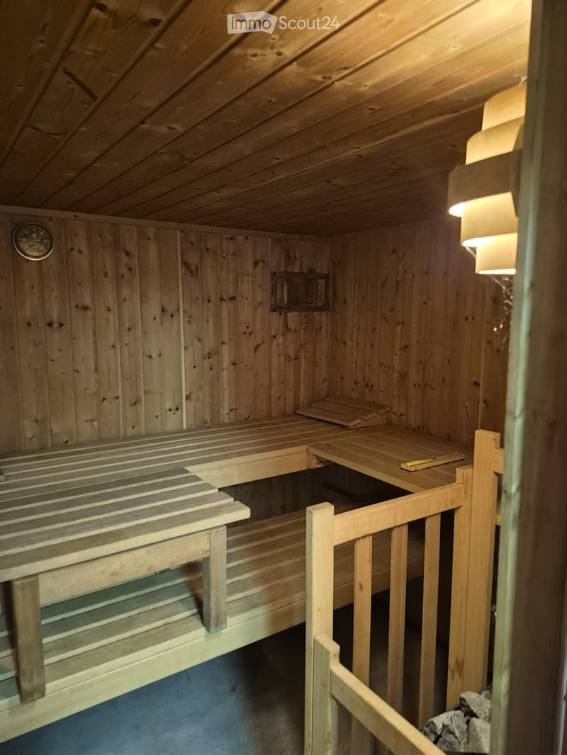 The image shows a wooden sauna with multiple wooden benches and a small wooden stool, illuminated by warm lighting. The sauna features a wooden ceiling and walls, with a small window and a thermometer visible on the wall.