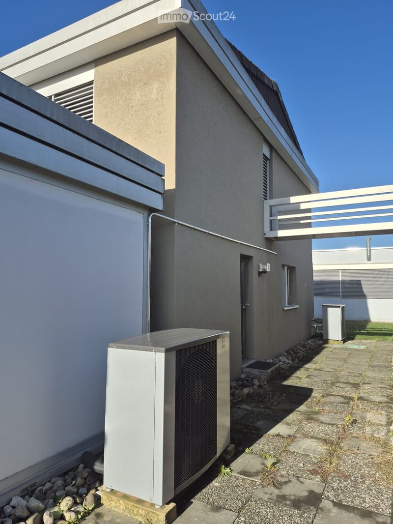 Modern two-story building with a flat roof, concrete facade, and a small balcony on the upper level. An air conditioning unit is positioned on the ground floor near the entrance.